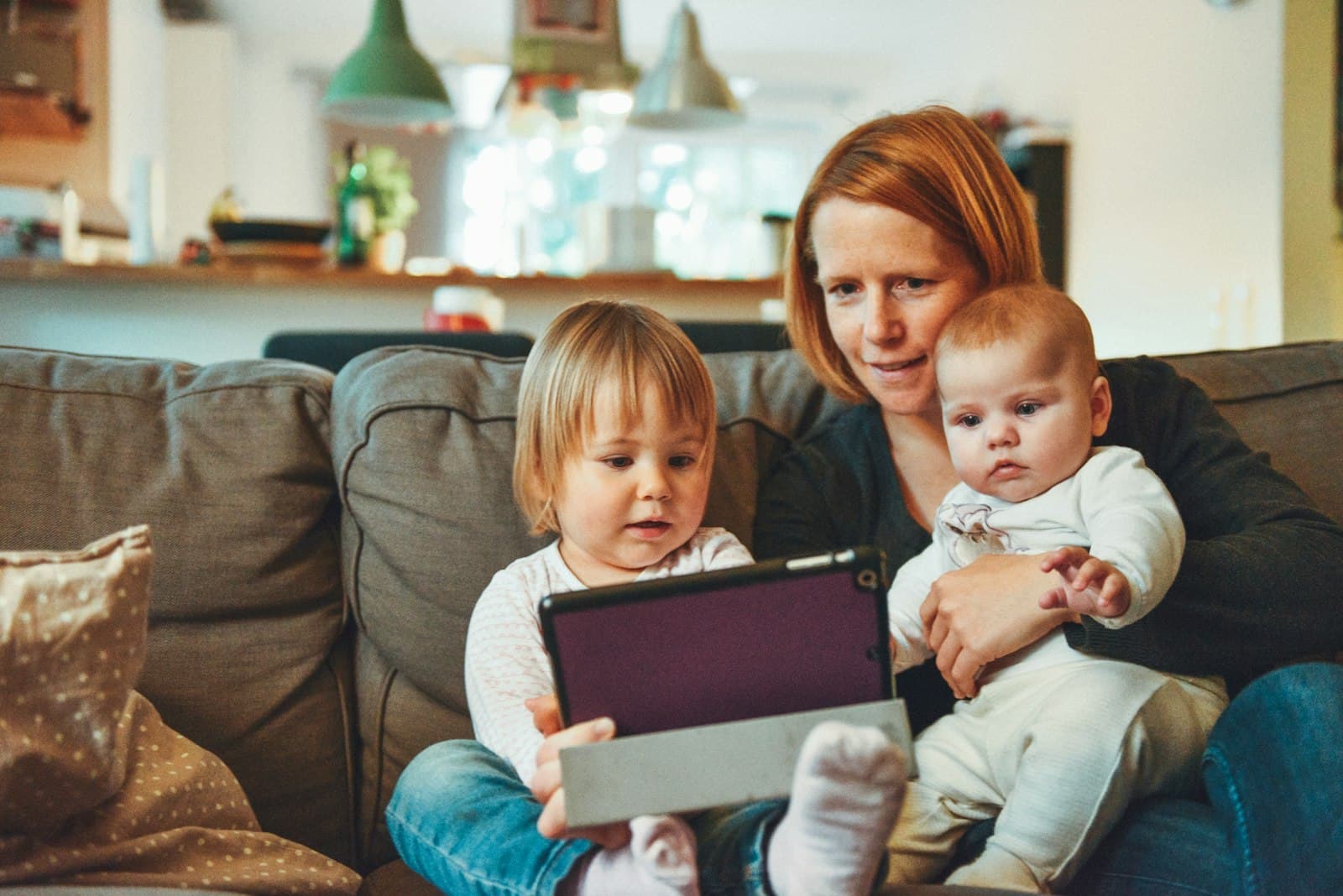 A parent and two young children looking at a tablet together on the sofa